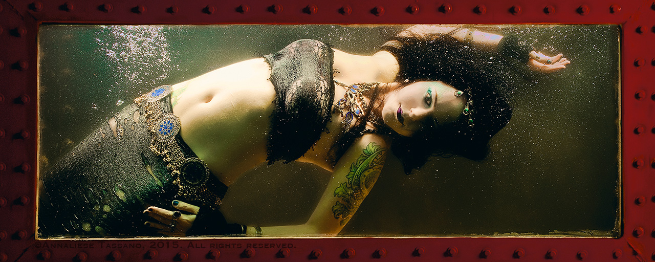 A tattooed, white mermaid with black hair and heavy silver jewelry floats in a bubble filled tank of water.