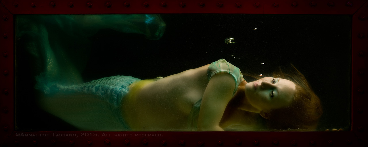 A pale, blond mermaid rests on the floor of the shadowy tank in which she is imprisoned.