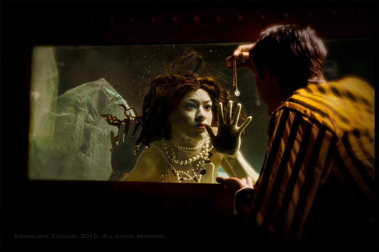 A mermaid in a tank is captivated and lured by the jewel a man in a yellow and black striped jacket outside holds up as bait to lure her.