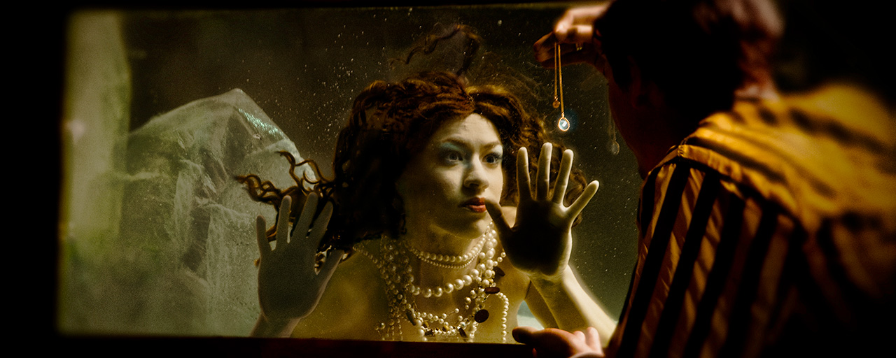 A mermaid in a tank is captivated and lured by the jewel a man in a yellow and black striped jacket outside holds up as bait to lure her.