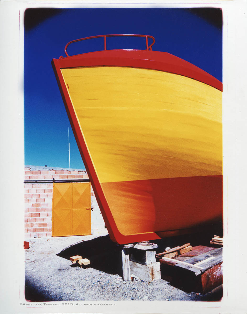 A cross process photo shows a finished wooden fishing boat painted brigth yellow, roange, and red. A brick wall with al large orange door and a a ricj blue sky in the background.