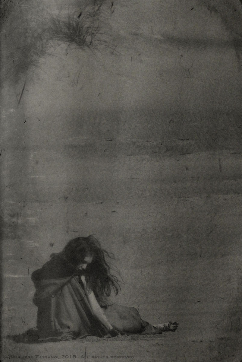 A young girl in a cloak sits on the beach playing in the sand in front of the dunes in the antique or vintage black and white photo.