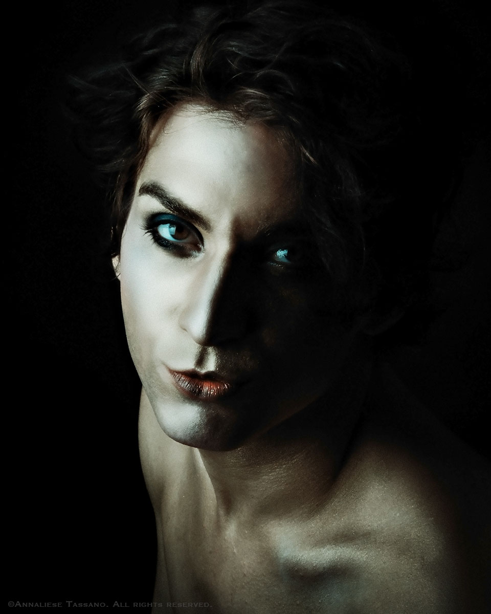 A handsome, young white man with theatrical make up looks at the camera from a dark background.