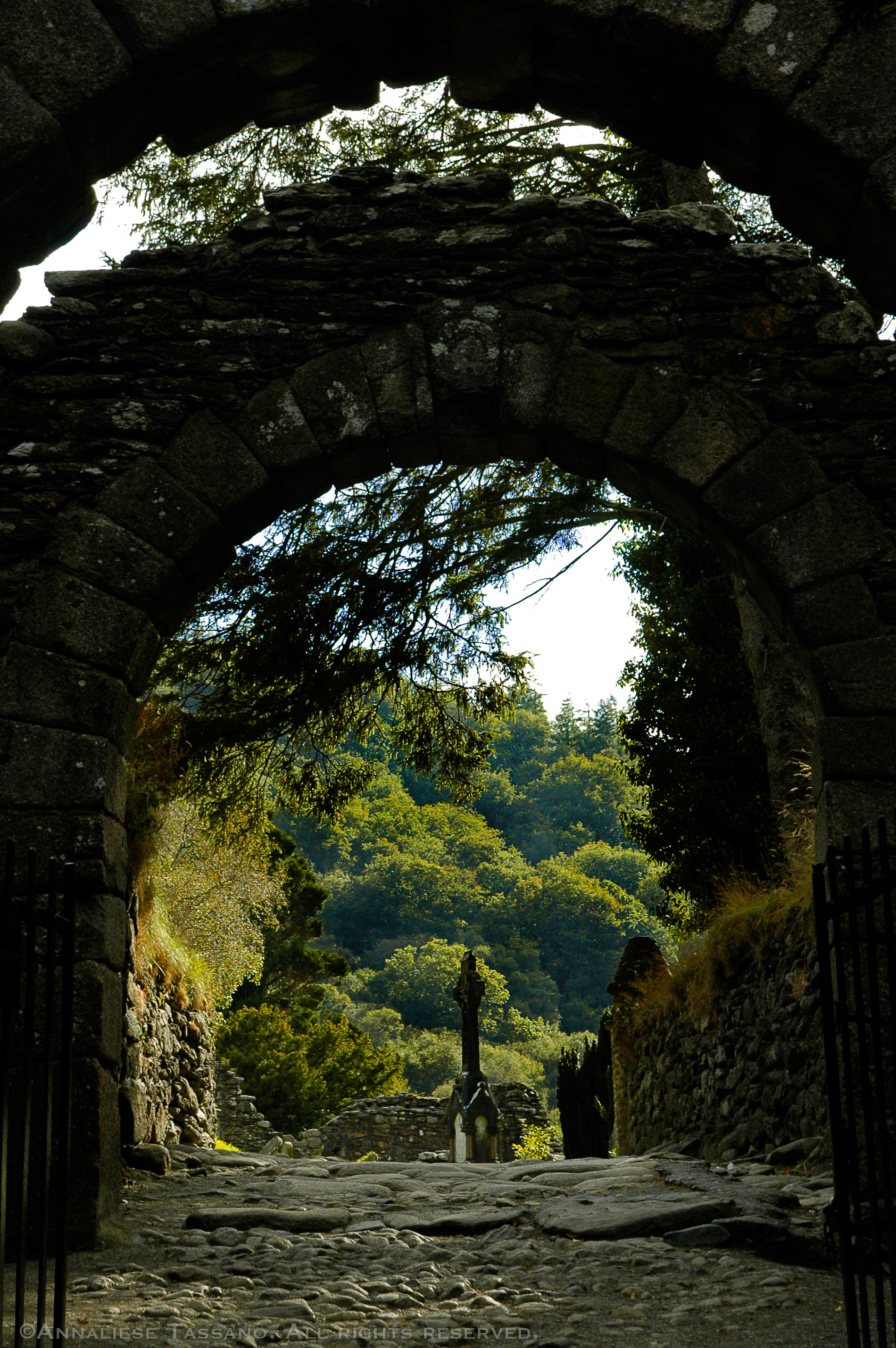 A hillside of green trees is seen in the sunshine beyond and ancient stone archway and cobblestone street in Glendalough, county Wicklow, Ireland.