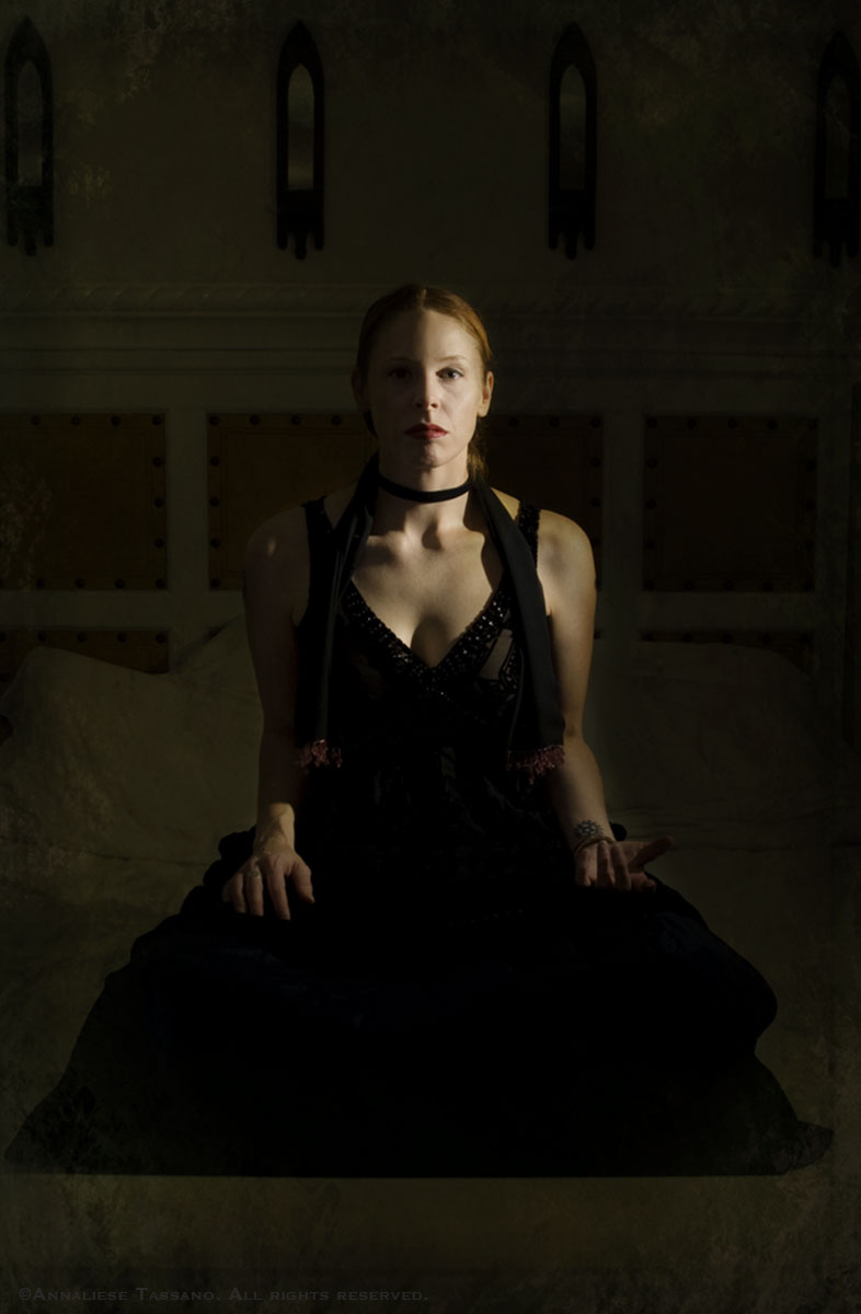 A white woman with red hair and lipstick wearing a black dress and choker sits crossed legs in a dark, wainscoted room, her left hand in a mudra.