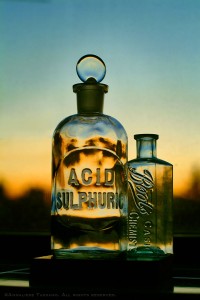 Antique glass bottles, on with a glass stopper, reading Acid Sulphuric and Boots Chemists cash, sit in a window as the sunset fades outside, in Brooklyn, New York.