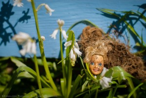 A dirty Barbie doll head, recently dug up from the garden is displayed in front of a bright blue house, nestled in greens and white flowers.