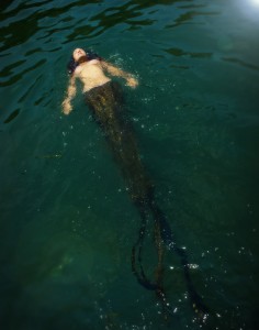 A beautiful young mermaid floats in green waters, sparkling in the sunlight.